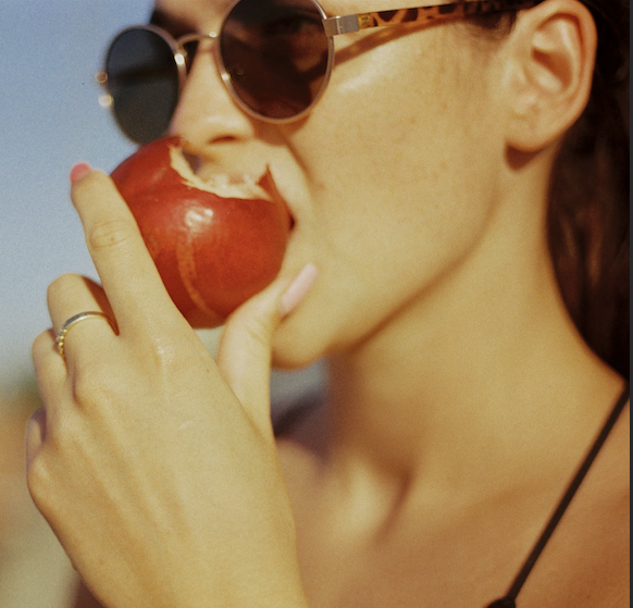 A woman with pink nails and a ring, wearing sunglasses, is about to bite into a red apple outdoors. Promote healthy habits with Eat more plants! (1-on-1 Health Coaching).